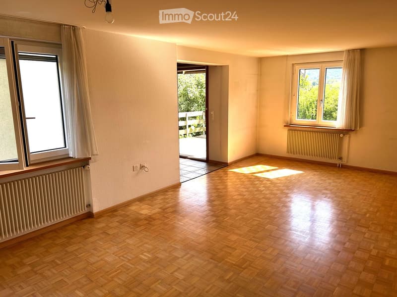 Empty room with wooden parquet flooring, large windows with curtains, radiators, and a door leading to a balcony or terrace area.