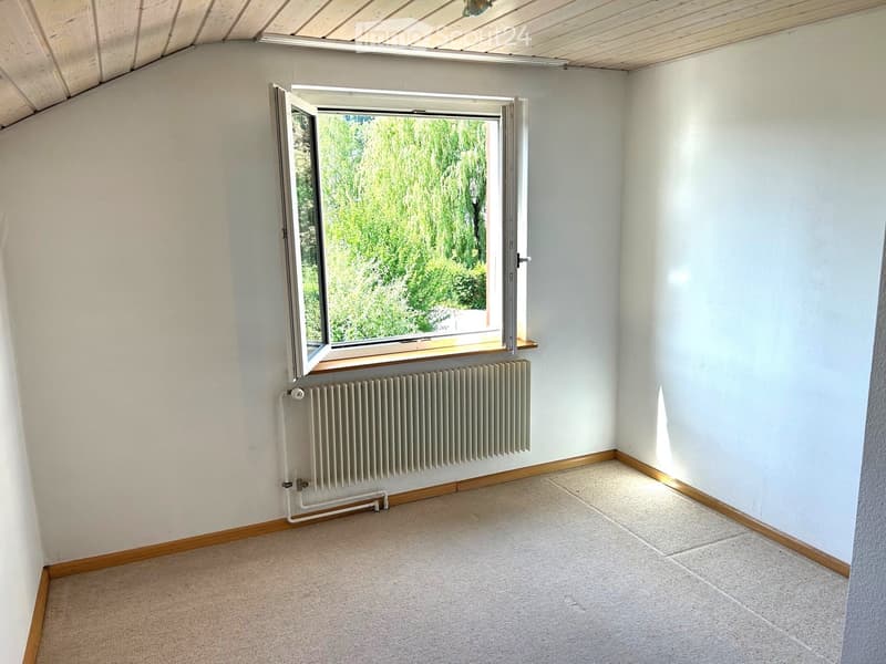 Empty room with white walls, wooden baseboards, carpeted floor, and a large window with a radiator below it.