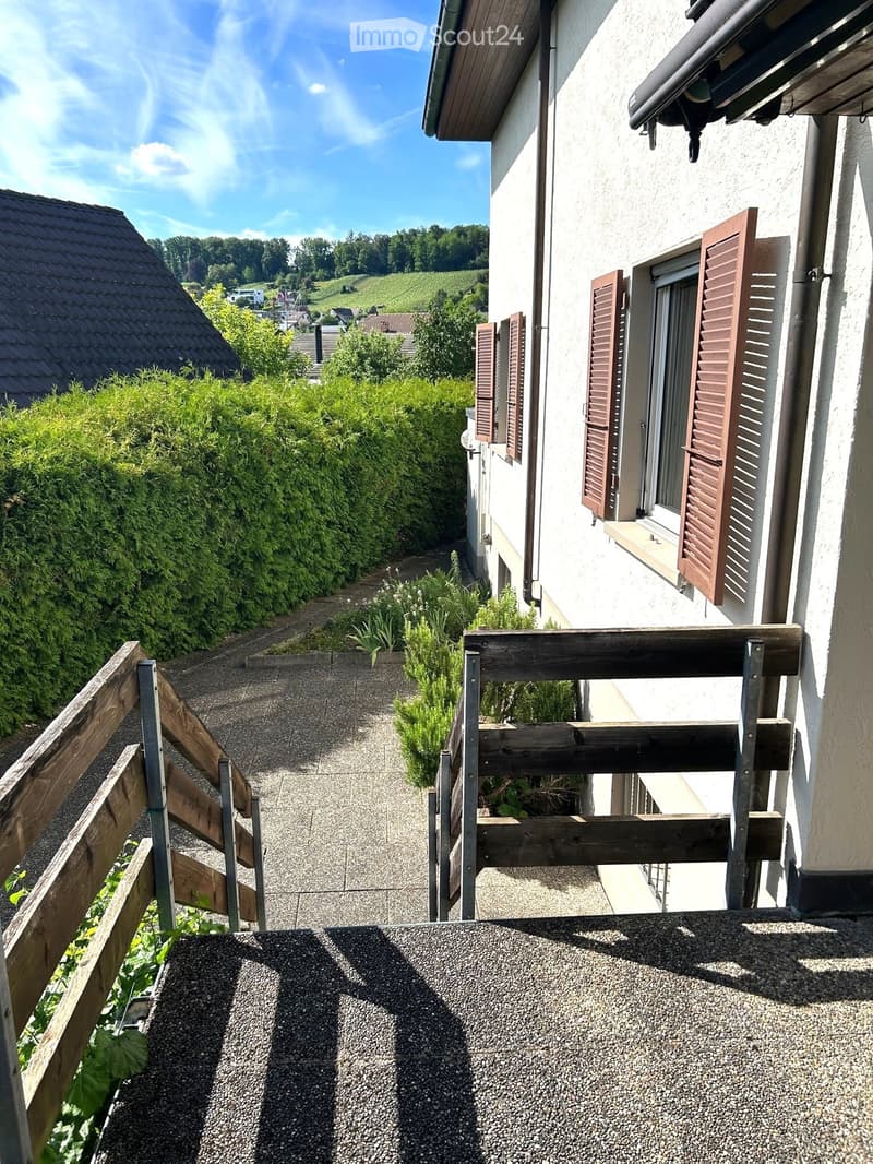 Balcony with wooden railing, gravel surface, and view of a house with shutters and surrounding greenery.