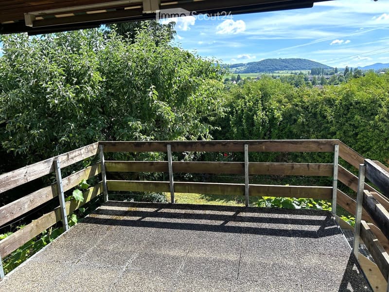 Wooden balcony with metal railings, overlooking a lush green landscape and distant hills.