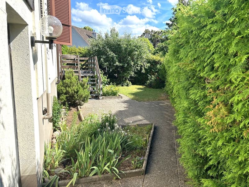 A narrow garden path lined with greenery and a wooden staircase leading to a raised area, with a wall-mounted light fixture on the left.