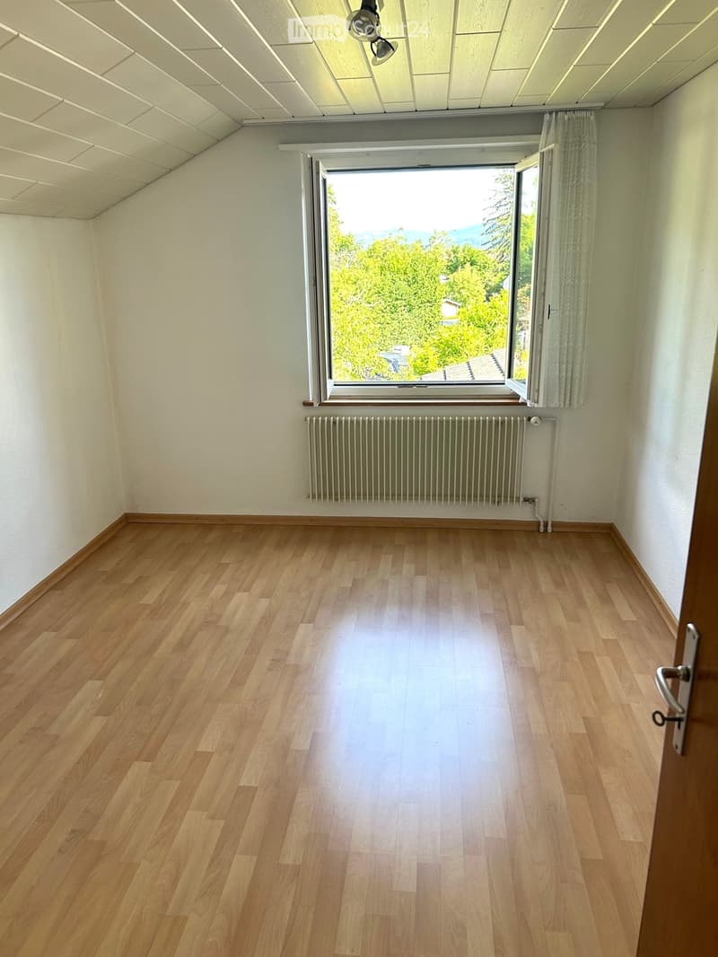 Empty room with light wooden flooring, white walls, a large window with a view of greenery, and a radiator below the window.