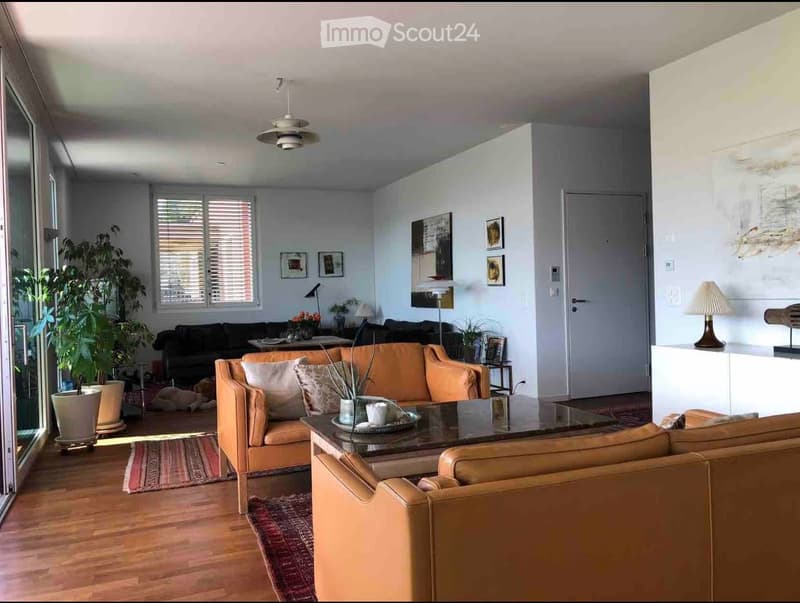 Modern living room with light-colored furniture, including a sofa, coffee table, and chairs. Features include large windows, a ceiling fan, and decorative plants.