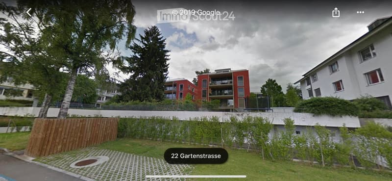 Modern multi-story residential building with red and white facades, surrounded by greenery and a paved pathway.