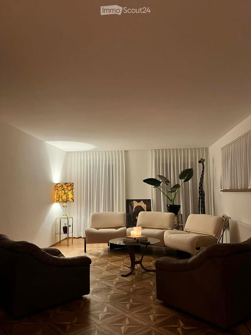 Modern living room with a cream-colored sectional sofa, a dark brown armchair, a glass coffee table, a decorative lamp, and a plant in a pot. The room features patterned tile flooring and large windows with white curtains.