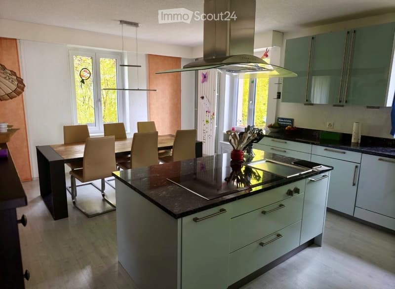 Modern kitchen with light green cabinets, dark granite countertops, a central island, and a dining area with a wooden table and beige chairs. Features include an induction cooktop, range hood, and pendant lighting.