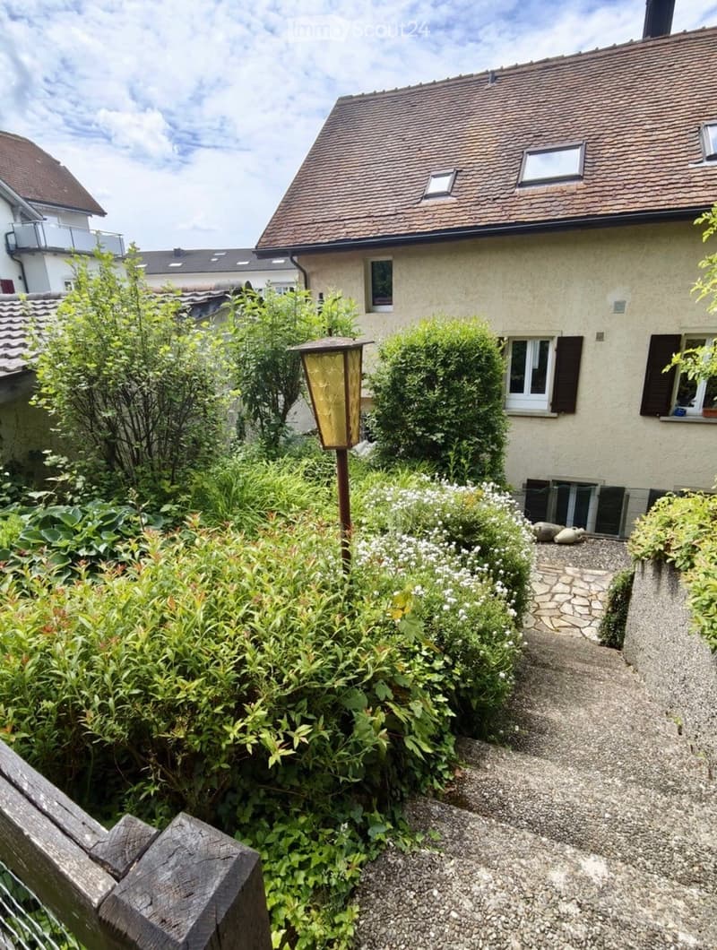 A well-maintained garden area with a pathway leading to a house, featuring various plants, shrubs, and a decorative lantern post.