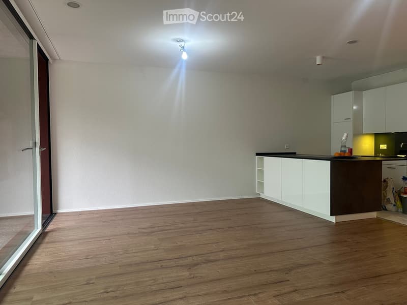 Empty kitchen with white cabinets, dark countertop, wooden flooring, and a large window with blinds.