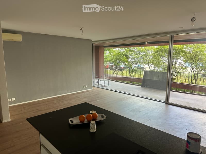 Modern kitchen with wooden flooring, black countertop, and large sliding glass doors leading to a balcony with outdoor furniture.