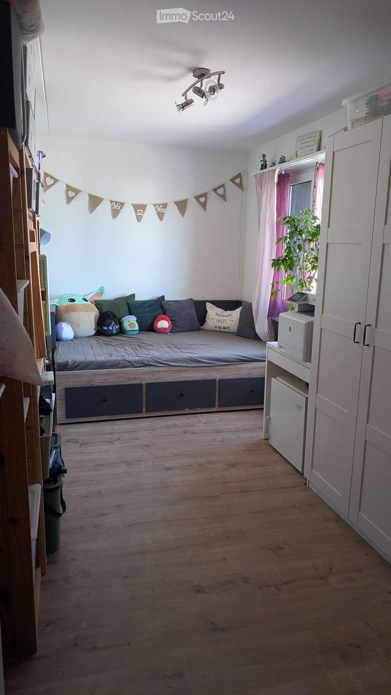 A cozy bedroom with a wooden floor, a built-in sofa bed with multiple pillows, a white cabinet, a potted plant near the window, and a small printer on a shelf.