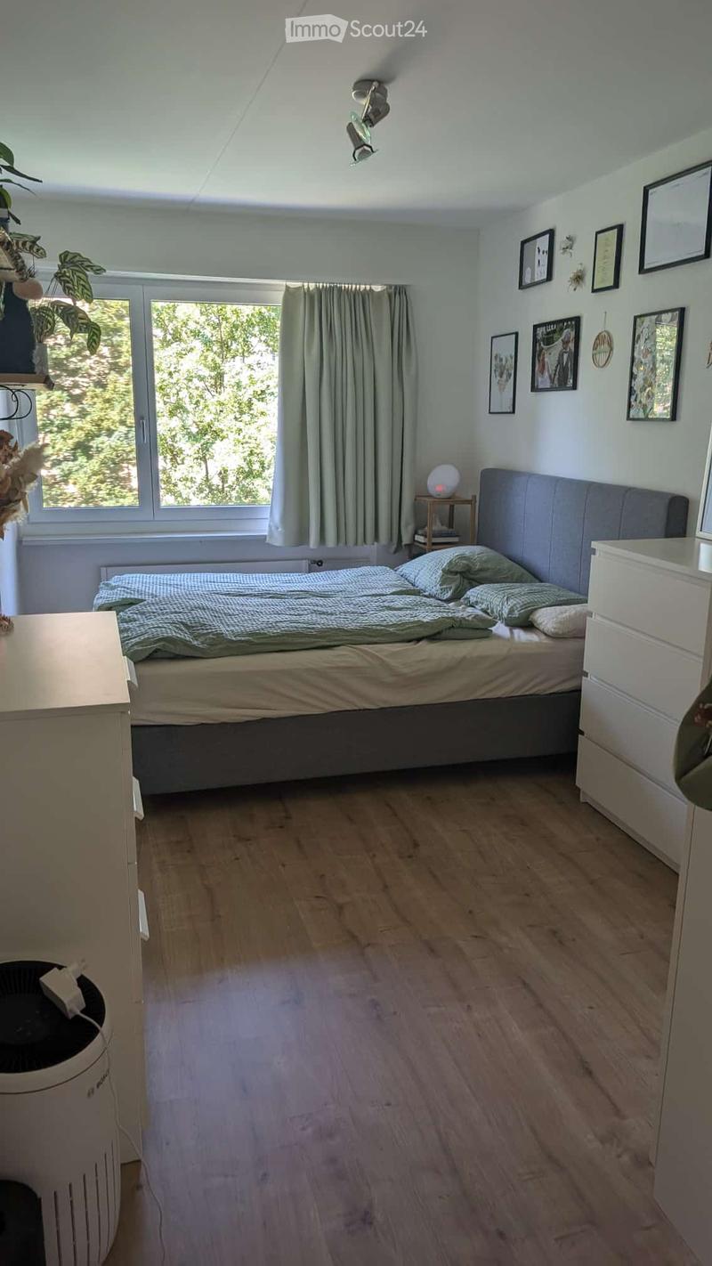 Cozy bedroom with wooden flooring, large window with green curtains, bed with grey headboard, nightstand with lamp, and decorative plants.