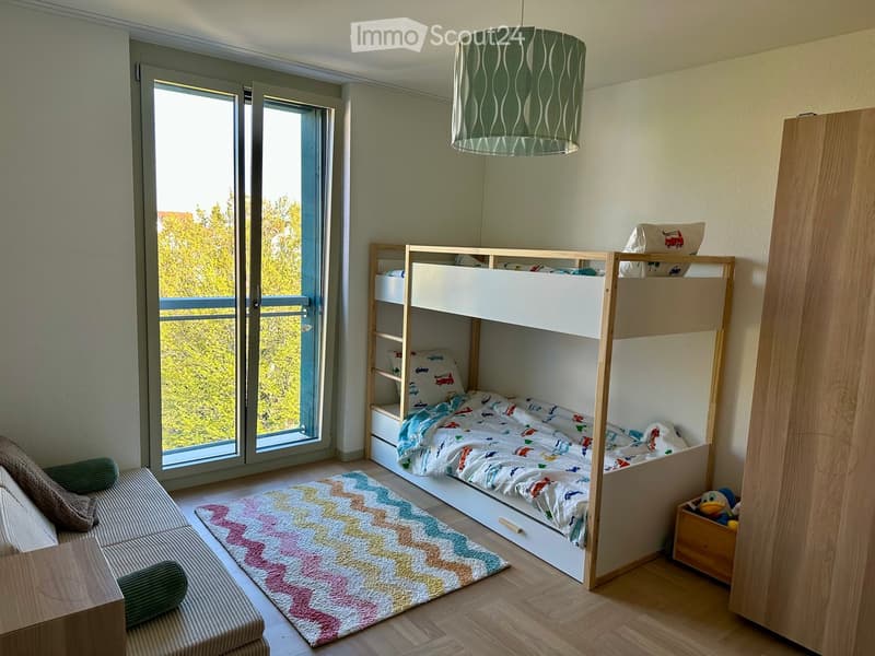 Bright bedroom with a bunk bed, colorful bedding, a sofa, a rug, and a view of greenery through large windows.