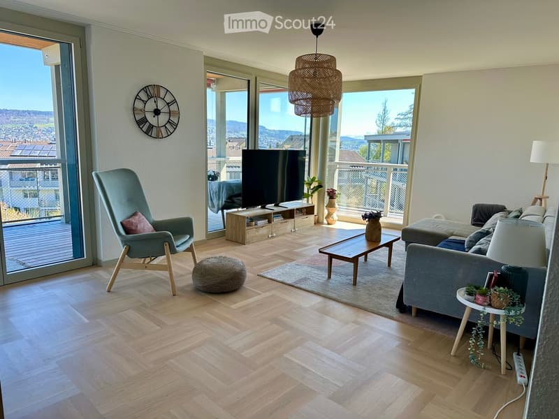 Modern living room with light wooden flooring, large windows offering a scenic view, a grey sofa, a wooden coffee table, a wicker hanging lamp, a TV, and decorative vases.