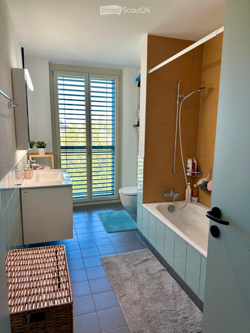 Modern bathroom with a bathtub, shower, toilet, sink, and a wicker basket. Features a large window with blinds and a small plant on the windowsill.