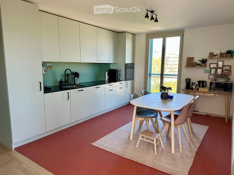 Modern kitchen with white cabinets, red tiled floor, and a dining area with a table and chairs. Features include a sink, oven, microwave, and coffee maker.