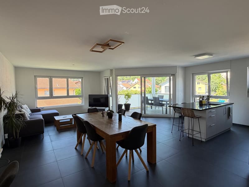 Open-plan living and dining area with a sofa, coffee table, dining table, chairs, kitchen island, and television. Large windows and sliding glass doors lead to a balcony with outdoor furniture.