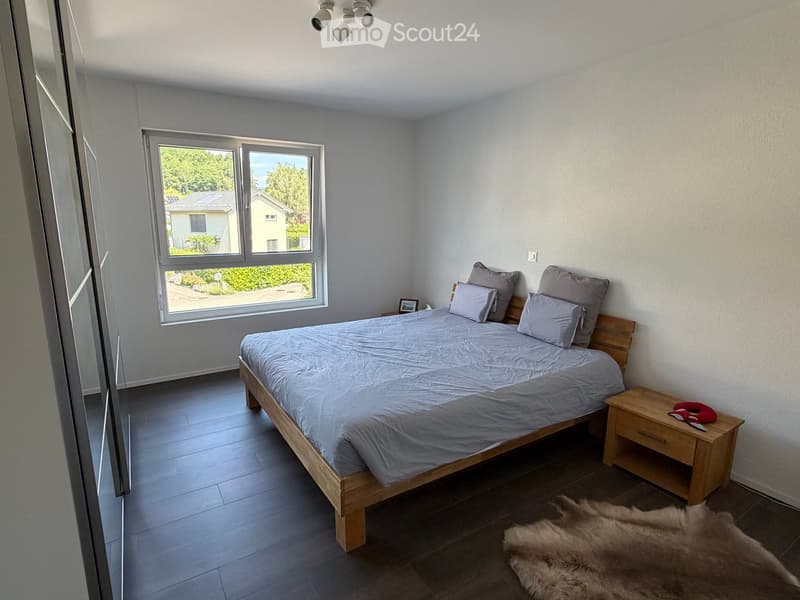 Modern bedroom with a wooden bed, grey bedding, nightstand, picture frame, and a furry rug on the floor.