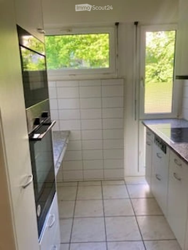 Modern kitchen with white tiled walls, white cabinets, a built-in oven, and a dishwasher. The space is clean and well-lit with natural light from the windows.