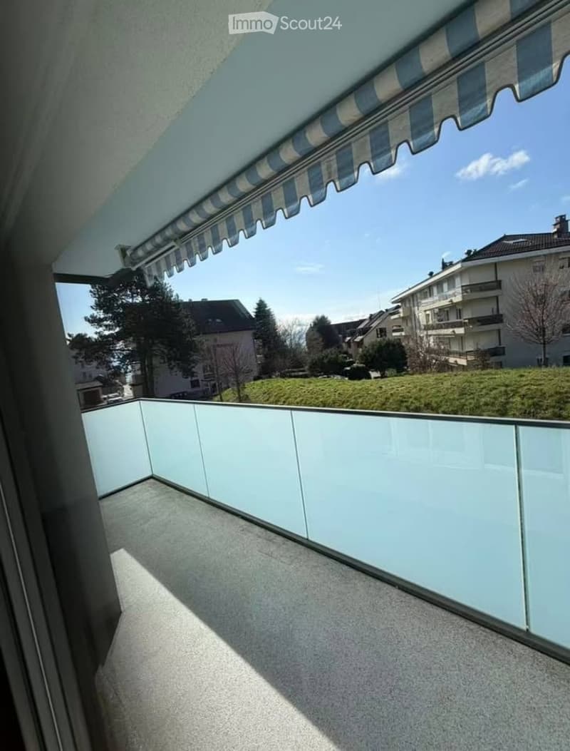 Balcony with a striped awning, glass railing, and a view of a green area with trees and buildings.