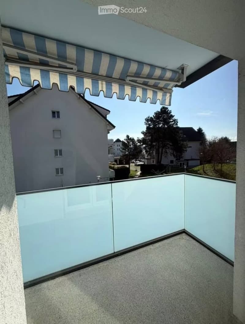 Balcony with glass railing, retractable awning, overlooking a residential area with houses and trees.