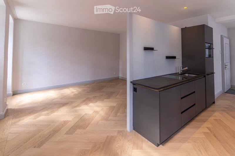 Modern kitchen with dark grey cabinets, a black countertop, and a double sink. The floor is made of light wooden parquet, and the walls are painted in a light grey color. The kitchen features built-in storage and a sleek, contemporary design.