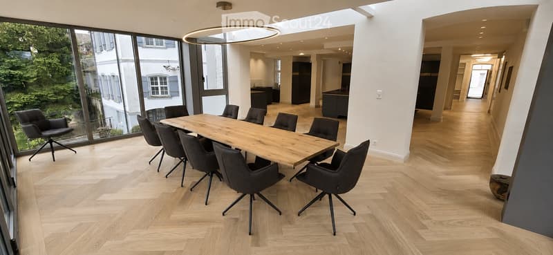Modern office or meeting room with a large wooden table surrounded by black chairs, large windows with a view of greenery, and a sleek hanging light fixture.