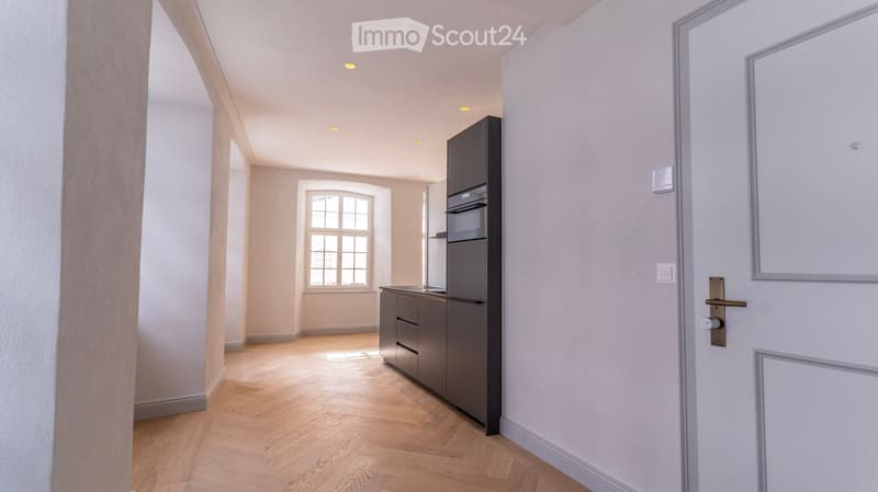 Modern kitchen with light wooden flooring, grey cabinets, and a built-in refrigerator. A large window allows natural light to fill the space.