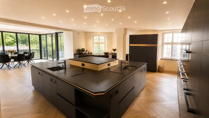 Modern kitchen with dark countertops, stainless steel sink, island, and built-in cabinets. Large windows and a dining area with chairs and a table are visible.