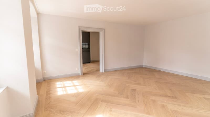 Empty room with light-colored walls and a herringbone-patterned wooden floor. A doorway leads to another room with a visible refrigerator.