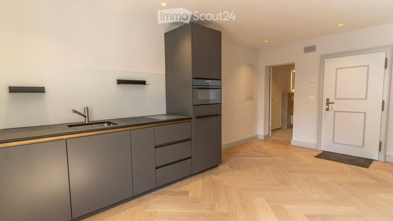 Modern kitchen with grey cabinets, a sink, faucet, and built-in oven. The floor is light wood, and the walls are white with open shelving.