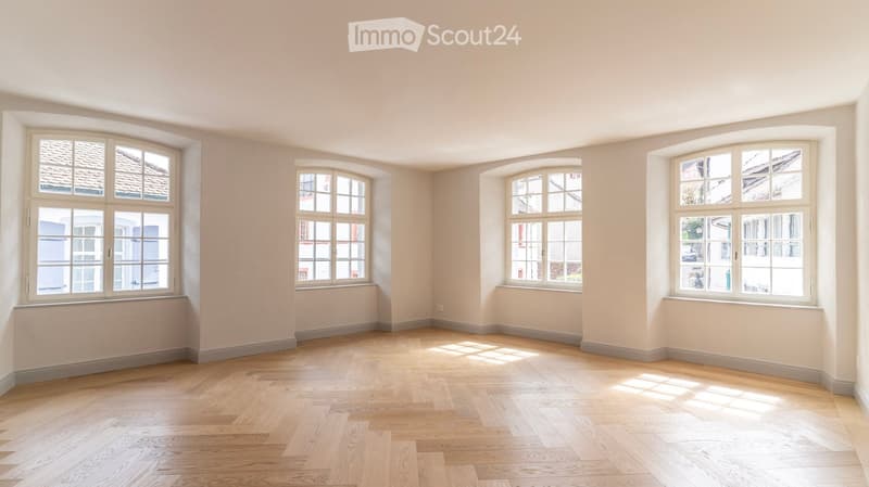 Empty room with light-colored walls and wooden flooring, featuring multiple large windows with white frames and frosted glass panes.