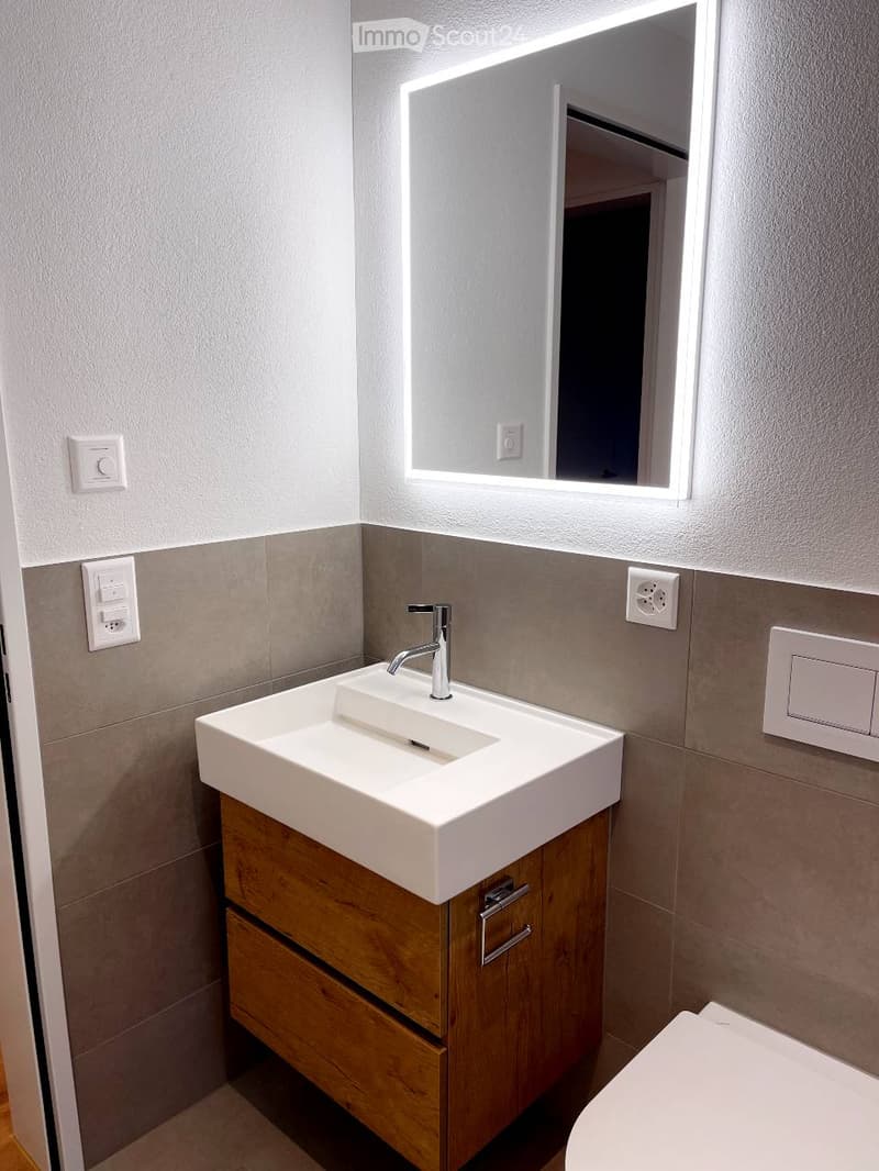 Modern bathroom with a white sink, wooden cabinet, and illuminated mirror above it. The walls are tiled in a neutral color scheme, and the faucet is sleek and contemporary.