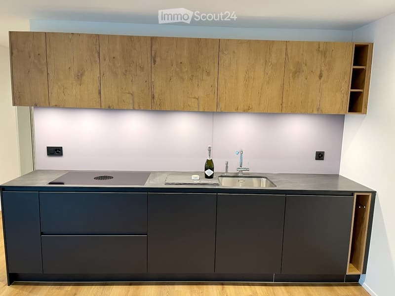 Modern kitchen with wooden upper cabinets, dark lower cabinets, and a stainless steel sink. A bottle is placed on the counter.