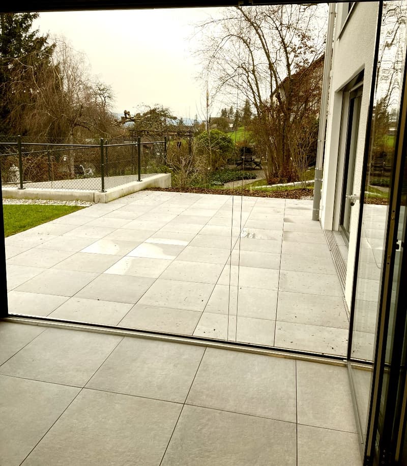 Modern balcony with large glass sliding doors, tiled floor, and a view of a landscaped garden with trees and a fence.