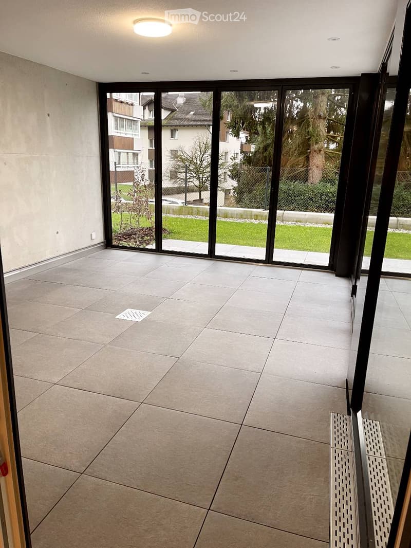 Modern hallway with large glass sliding doors leading to a green outdoor area, tiled floor, and recessed ceiling lighting.