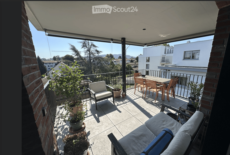 Balcony with a sofa, chairs, a table, and several potted plants, overlooking a cityscape with buildings and trees.