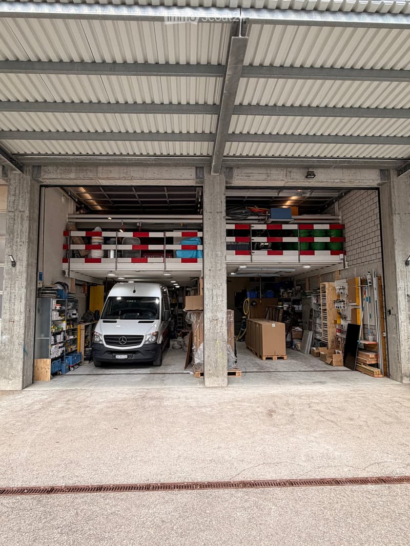 Large, open garage with concrete floors and walls, featuring a parked van, shelves with storage items, and various tools and equipment.