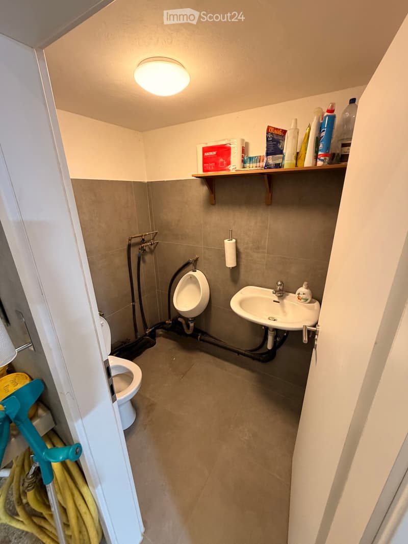 Small bathroom with beige tiled walls and floor, featuring a toilet, urinal, sink, and a wooden shelf holding cleaning supplies and toilet paper.