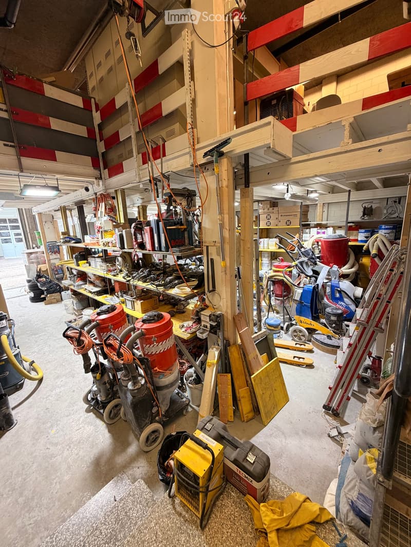 A cluttered garage filled with various tools and equipment, including vacuum cleaners, a saw, a drill, a ladder, and a toolbox. Shelves and cabinets hold additional items, and wooden planks are scattered around.