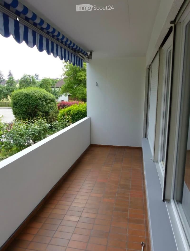 Balcony with tiled floor, white walls, and striped awning, overlooking a green garden area.