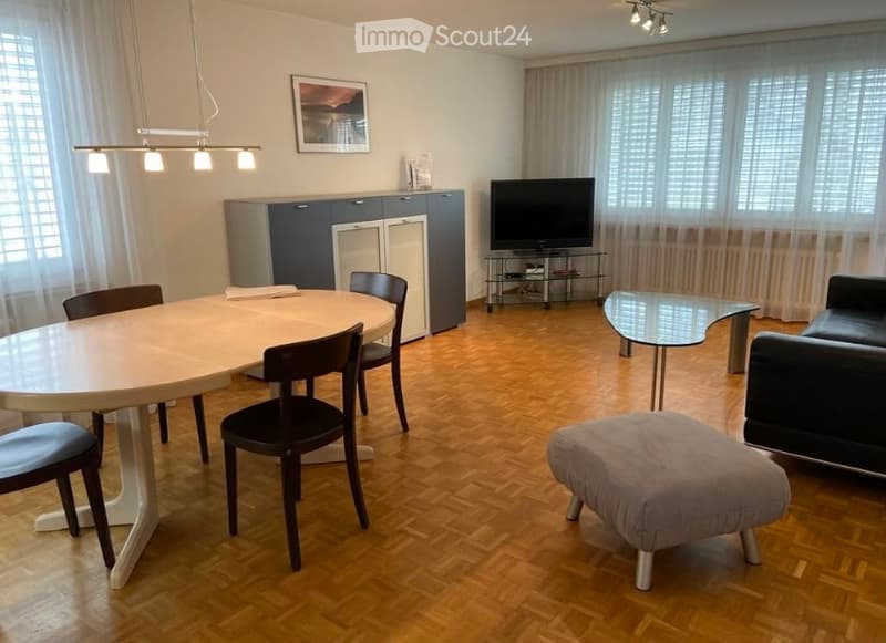 Modern furnished room with hardwood flooring, a round dining table with chairs, a sofa with an ottoman, a television on a stand, and a cabinet with a picture frame on top.