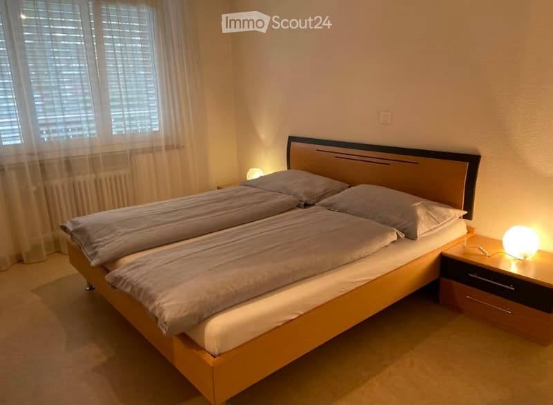 A modern bedroom with a double bed, two pillows, a nightstand with a lamp, and a window with blinds and curtains.