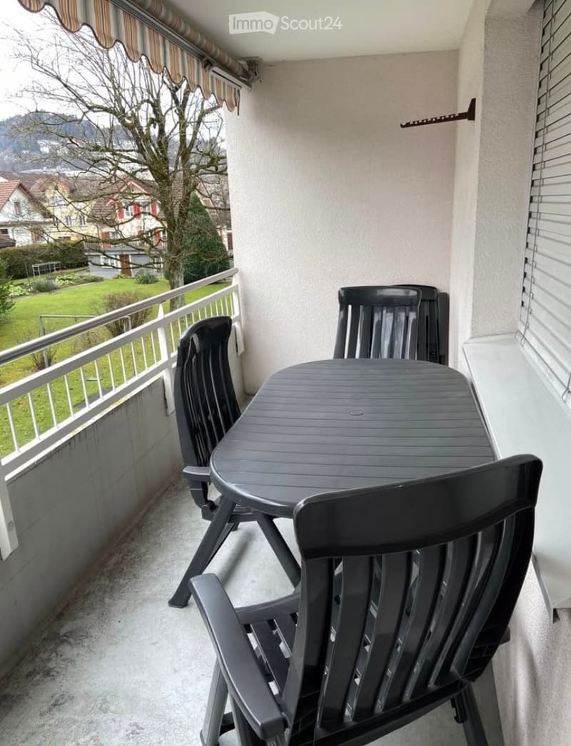 Balcony with a table and chairs, overlooking a green landscape with houses and trees.