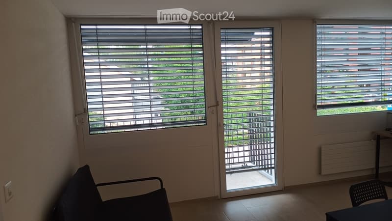 Empty room with wooden flooring, large windows with blinds, and a sliding glass door leading to a balcony.