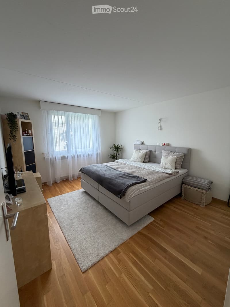 Modern bedroom with wooden flooring, large bed with grey bedding, nightstand, small stool, shelf with decorative items, and a window with sheer curtains.