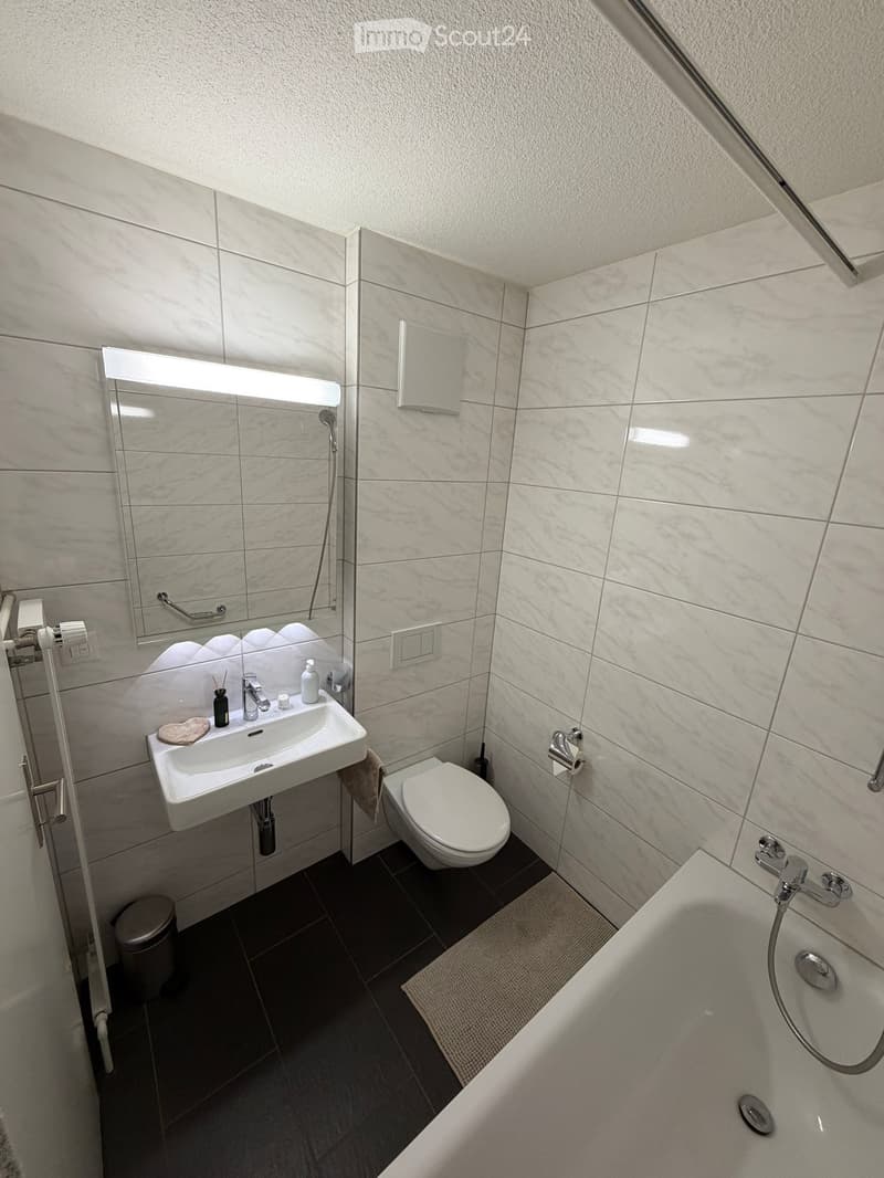 Modern bathroom with white tiles, a sink, toilet, bathtub, and a large mirror with built-in lighting. Includes a towel and a small trash can.