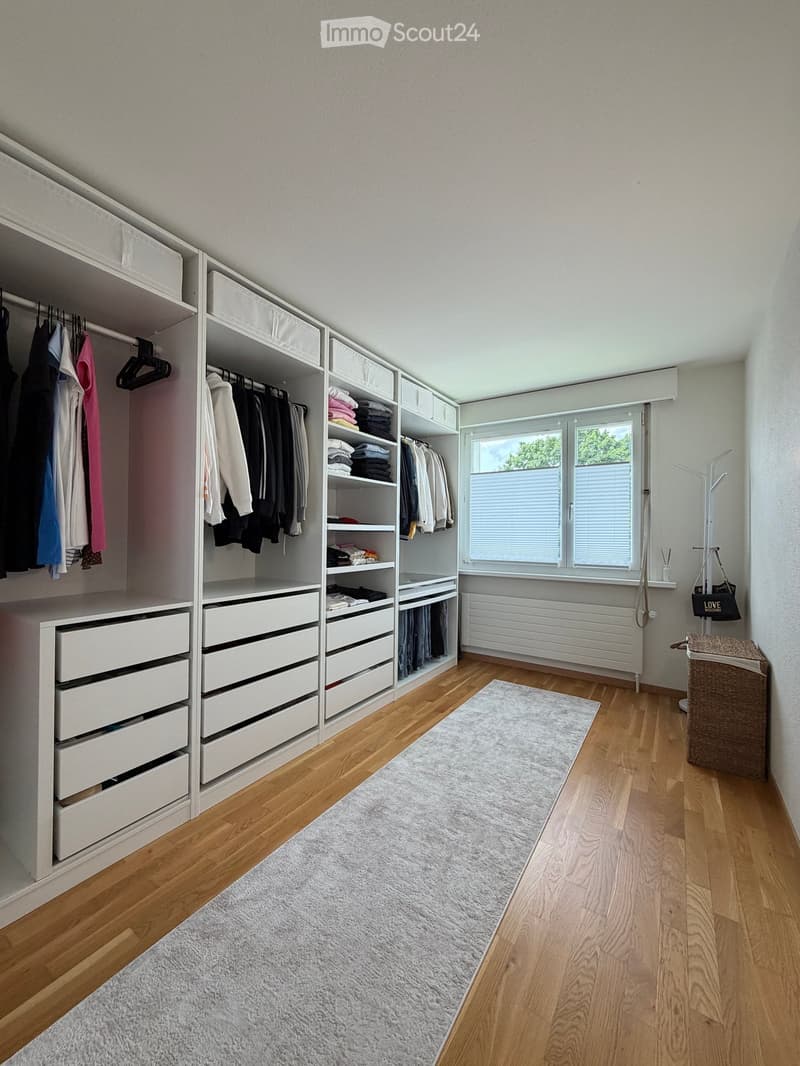 A well-organized walk-in closet with white built-in wardrobes, multiple drawers, and a variety of neatly arranged clothing items. The room features wooden flooring, a gray rug, and a window with blinds.