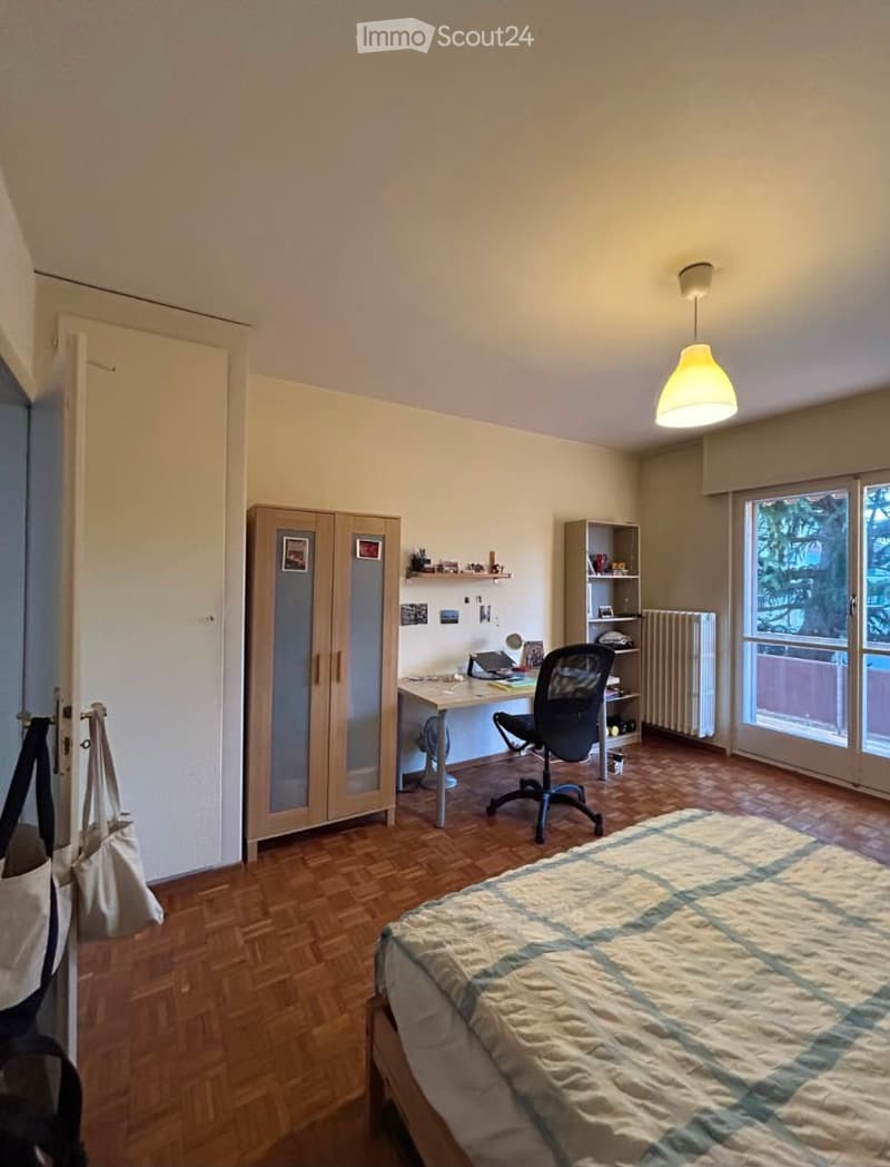 A well-lit bedroom with a wooden floor, a bed with a striped duvet, a desk with a chair, a wardrobe, and a shelf with various items.