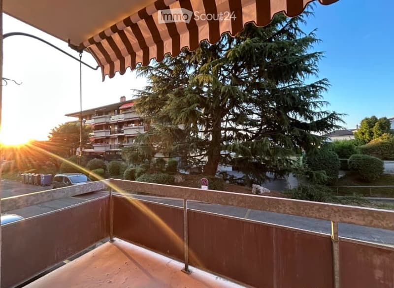 Balcony with striped awning, overlooking a tree-lined street and multi-story apartment building, with sunset visible.