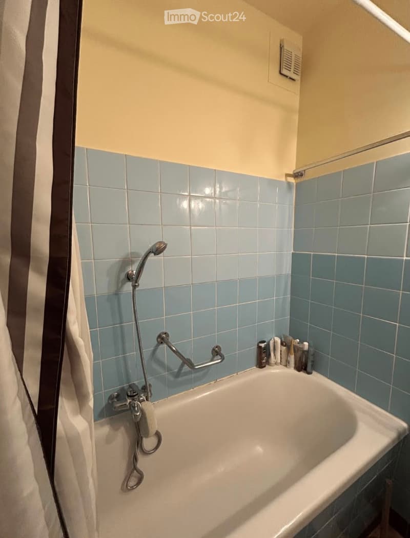 Bathroom with a white bathtub, blue tiled walls, a showerhead, a handrail, and a towel on the side.